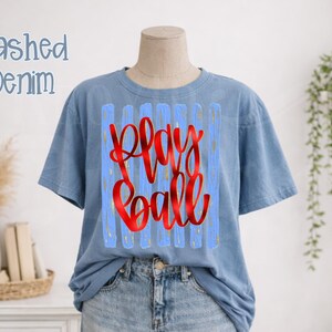 May include: A light blue t-shirt with the words "Play Ball" in red script. The text is set against a light blue background with vertical stripes. The shirt is displayed on a mannequin.