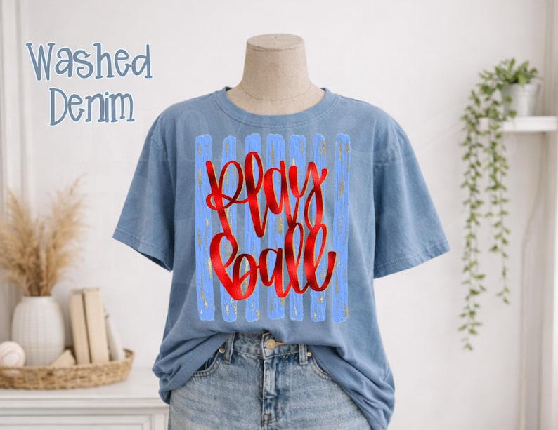 May include: A light blue t-shirt with the words "Play Ball" in red script. The text is set against a light blue background with vertical stripes. The shirt is displayed on a mannequin.