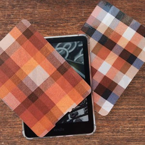 May include: Kindle e-reader in a clear case, with two square plaid coasters. The coasters have a pattern of orange, brown, white, and blue squares. The e-reader is on a wooden surface.