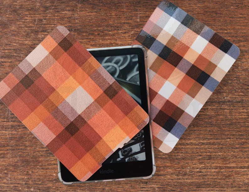 May include: Kindle e-reader in a clear case, with two square plaid coasters. The coasters have a pattern of orange, brown, white, and blue squares. The e-reader is on a wooden surface.