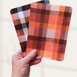 May include: Two rectangular plaid coasters are held in a hand. One coaster features a pattern of orange, red, and brown squares. The other coaster has a pattern of brown, white, and blue squares. The coasters have rounded corners.