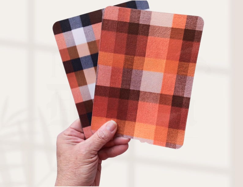 May include: Two rectangular plaid coasters are held in a hand. One coaster features a pattern of orange, red, and brown squares. The other coaster has a pattern of brown, white, and blue squares. The coasters have rounded corners.