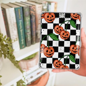 May include: A clear phone case featuring a black and white checkered pattern with orange jack-o'-lantern cherry designs. The case has a transparent border and is held in a person's hand. The design includes green leaves and stems.