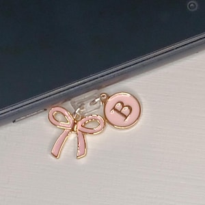 May include: A pink and gold charm with a bow and a circle with the letter "B" in the center. The charm is attached to a black smartphone.