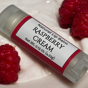 May include: A clear tube of natural lip balm with a red label that reads "Natural Lip Balm Raspberry Cream Net Wt. 0.15 oz. (4.25g)". The tube is sitting on a white surface with two red raspberries.
