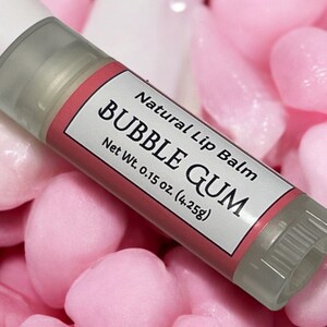 May include: A clear tube of lip balm with a pink label that reads "Natural Lip Balm Bubble Gum" and "Net Wt. 0.15 oz. (4.25g)". The tube is surrounded by pink heart-shaped candies.