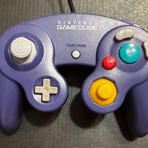 May include: A purple Nintendo GameCube video game controller featuring a white analog stick, a white D-pad, a yellow C-stick, a large green A button, a red B button, gray X and Y buttons, and a central white Start/Pause button.