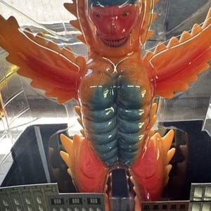 Puede incluir: Una figura de acción de monstruo de color naranja y rojo brillante con el pecho azulado y alas grandes, encerrada en un empaque de plástico transparente. La figura se coloca sobre una base de cartón impresa con un gráfico de edificios urbanos.