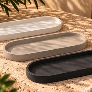 May include: Three oval-shaped trays with raised edges, arranged in a staggered formation. The trays are made of a solid material in three distinct colors: a light off-white, a medium gray, and a dark charcoal black, each with a smooth matte finish.