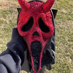 May include: A red and black Scream mask, embellished with sparkling red and black rhinestones. The mask has black eye holes and a black mouth. The mask has two red horns on top. The mask is held up by a black hood.