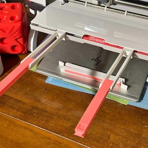 May include: A Cricut cutting machine with a white and red frame, and a silver cutting surface. Two pink and white support arms extend from the machine. A red, cube-shaped object and a white and red object are on the left.