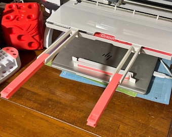 May include: A Cricut cutting machine with a white and red frame, and a silver cutting surface. Two pink and white support arms extend from the machine. A red, cube-shaped object and a white and red object are on the left.