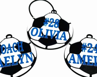 Puede incluir: Tres adornos con forma de balón de fútbol con paneles en blanco y negro. Cada adorno tiene un nombre y un número azules: "#28 OLIVIA", "COACH DAELYN" y "#24 AMELIA". Cada uno tiene un pequeño lazo negro en la parte superior.