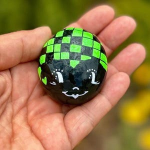 May include: A smooth, round black stone painted with a bright green checkered pattern on the top half. The lower half features a hand-painted white face with closed eyes, long eyelashes, and a small heart-shaped mouth, plus a small green heart.