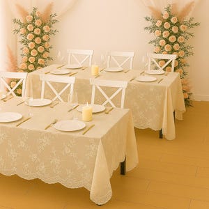 May include: A dining table setting with a cream-colored floral lace tablecloth, white plates, and gold-colored cutlery. White chairs are arranged around the table, with candles and wine glasses adding to the ambiance. Floral arrangements decorate the background.