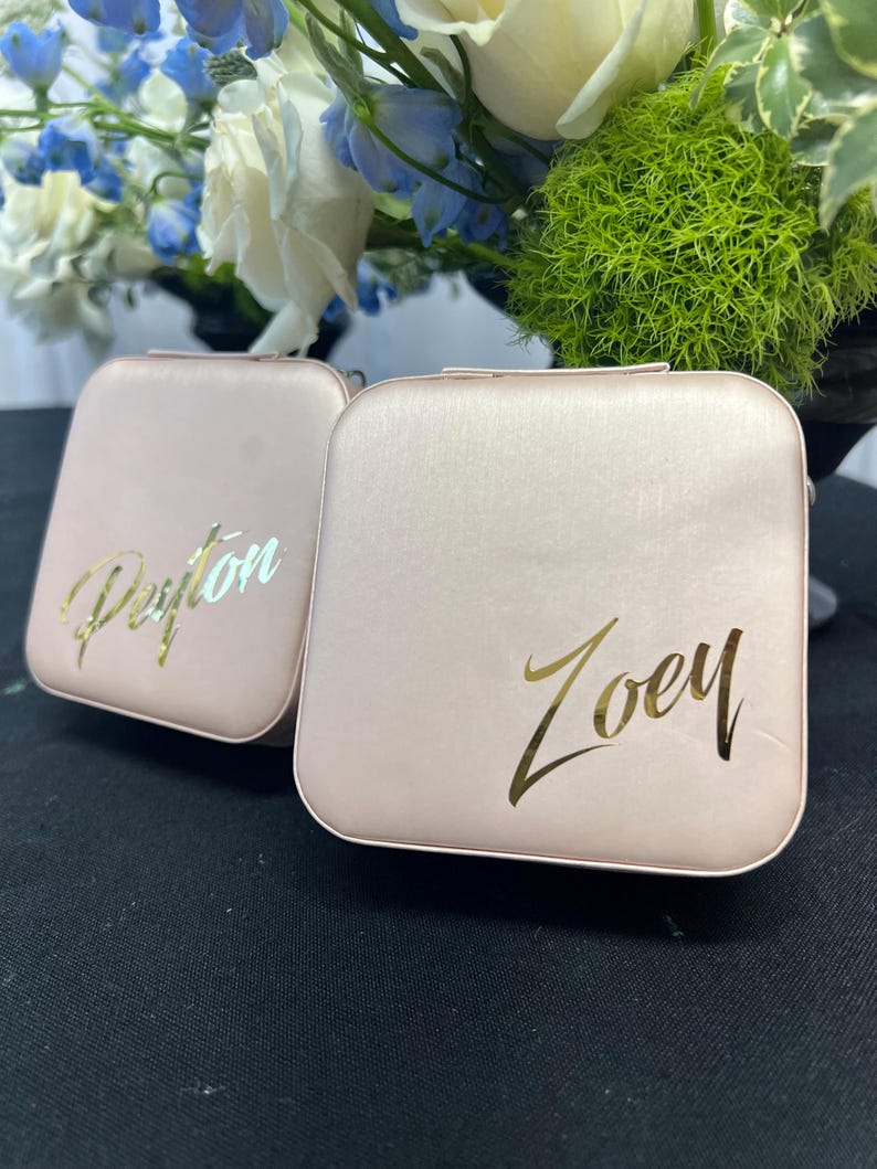 Pode incluir: Duas pequenas caixas quadradas cor-de-rosa com letras douradas. A caixa &agrave; esquerda diz "Peyton" e a caixa &agrave; direita diz "Zoey".