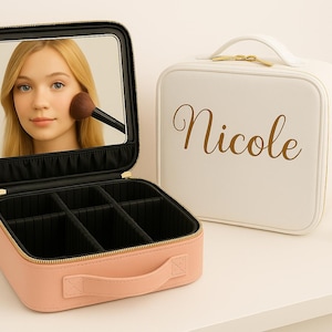 May include: A pink makeup case with a mirror, open to reveal compartments. A white case with "Nicole" in gold script is nearby. Lipstick and a gold tube are on the table. The mirror reflects a person applying makeup.