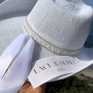 May include: White straw hat with a white pearl and rhinestone trim. The hat has a white ribbon tied around it. The hat has a tag that says "Lace & Honey atelier".