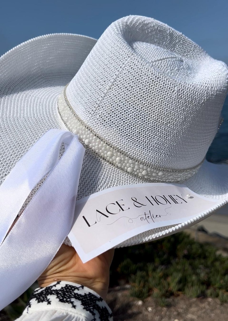 May include: White straw hat with a white pearl and rhinestone trim. The hat has a white ribbon tied around it. The hat has a tag that says "Lace & Honey atelier".