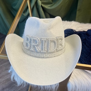 May include: A white cowboy hat featuring a textured woven finish and a wide brim. The front of the hat is adorned with the word BRIDE in large, silver-toned block letters outlined with small white pearls and a matching beaded hat band.