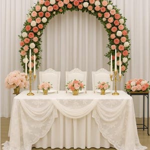 May include: A wedding reception table is set with a white tablecloth and three white chairs. A large floral arch of pink and white roses frames the table. Gold candle holders and small floral arrangements decorate the table.