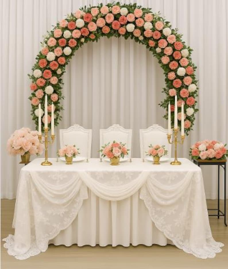 May include: A wedding reception table is set with a white tablecloth and three white chairs. A large floral arch of pink and white roses frames the table. Gold candle holders and small floral arrangements decorate the table.