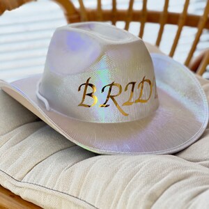 May include: A white iridescent cowboy hat with the word "Bride" written in gold letters.