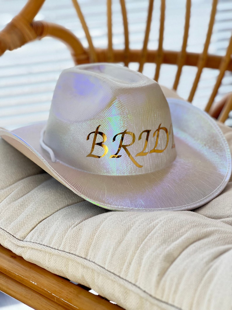 May include: A white iridescent cowboy hat with the word "Bride" written in gold letters.
