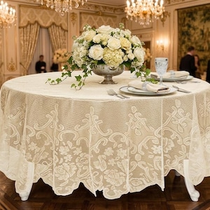 May include: A round, cream-colored lace tablecloth covers a table set for a formal event. A floral centerpiece of white roses and greenery sits in a silver vase. The table is set with plates, silverware, and glassware. The room has chandeliers and ornate decor.