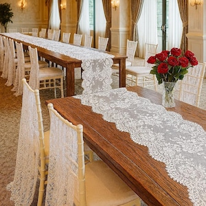May include: A formal dining room setup with long wooden tables adorned with white lace table runners. Chairs are draped with matching lace. A vase of red roses sits on one table. The room has large windows and elegant decor.