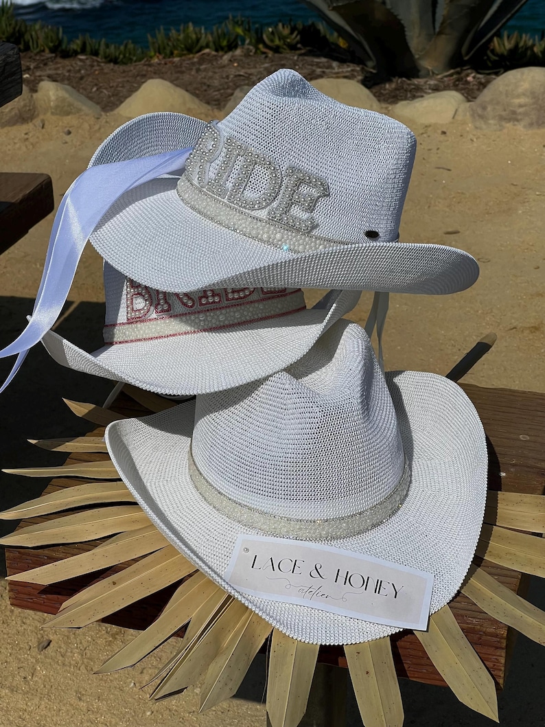 May include: Three white cowboy hats with a beaded trim. The top hat has the word "Bride" spelled out in rhinestones. The bottom hat has a tag that says "Lace & Honey Atelier".