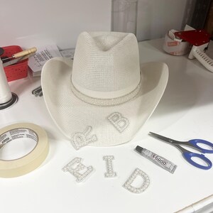 May include: A white cowboy hat with a pearl trim and the word "BRID" spelled out in pearl letters. The letters are on a white surface with a roll of tape, a tube of glue, and a pair of scissors.