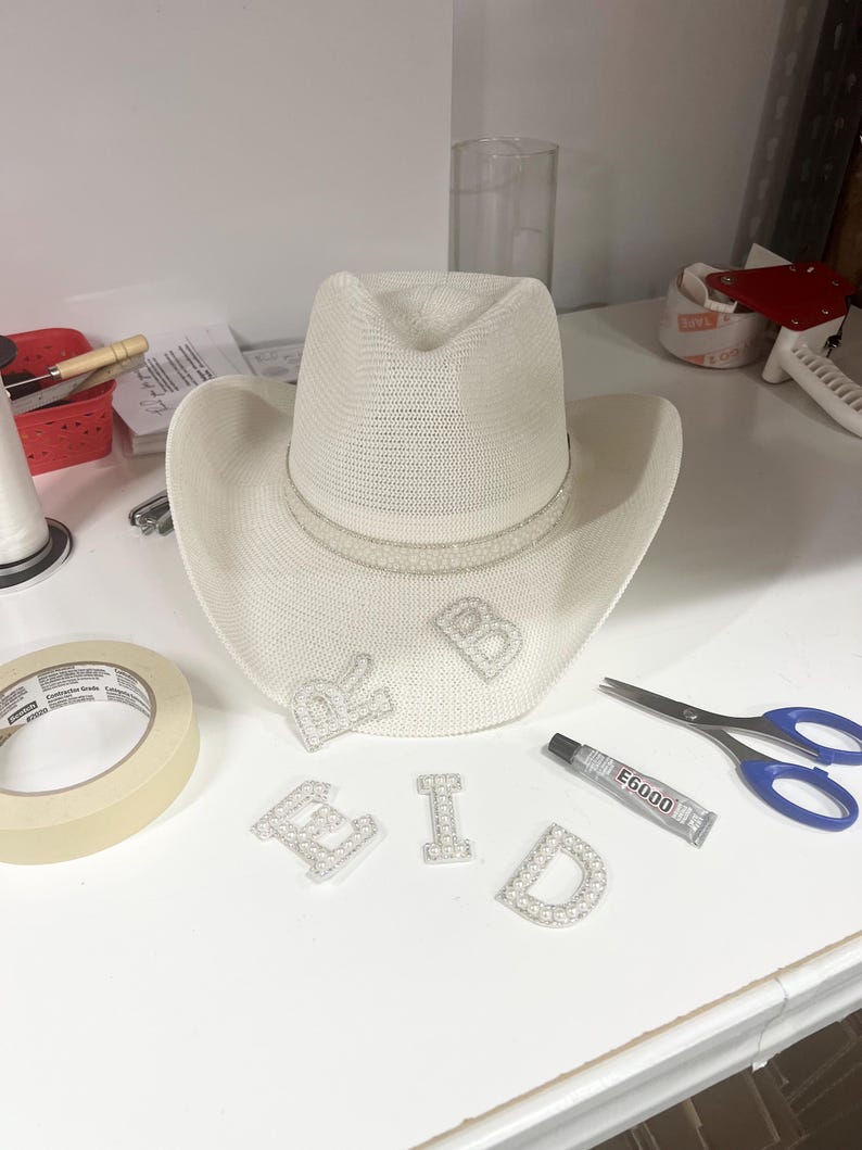 May include: A white cowboy hat with a pearl trim and the word "BRID" spelled out in pearl letters. The letters are on a white surface with a roll of tape, a tube of glue, and a pair of scissors.
