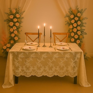 May include: A romantic table setting with a cream-colored lace tablecloth, two wooden chairs, and lit candles in brass holders. The table is set for two with plates, silverware, and wine glasses. Floral arrangements of peach roses and greenery flank the table.
