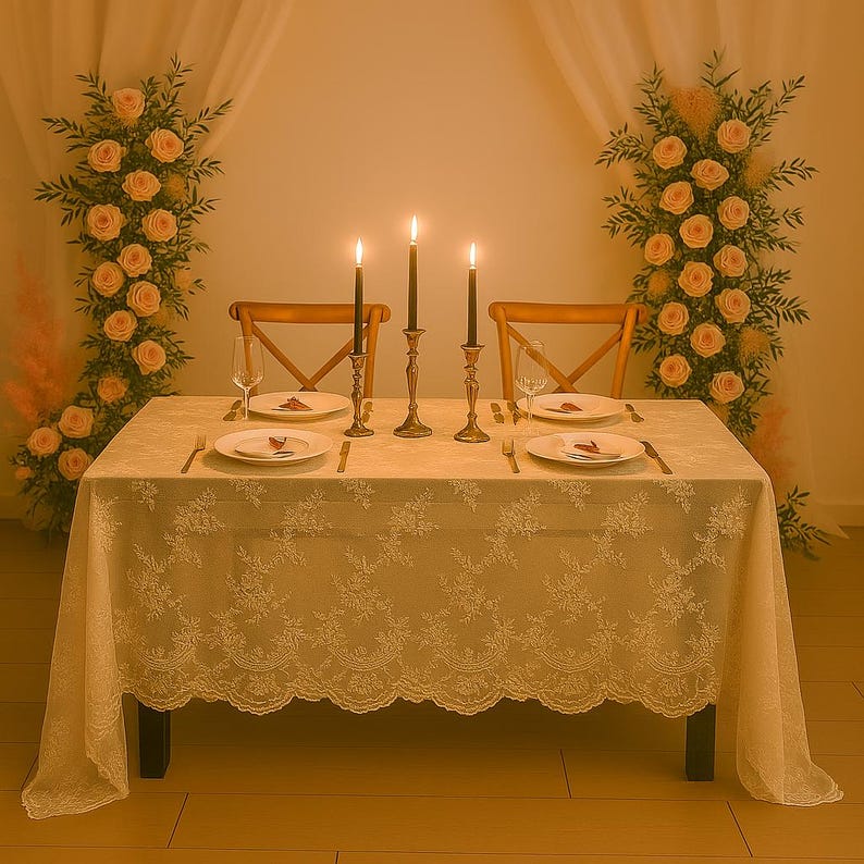 May include: A romantic table setting with a cream-colored lace tablecloth, two wooden chairs, and lit candles in brass holders. The table is set for two with plates, silverware, and wine glasses. Floral arrangements of peach roses and greenery flank the table.