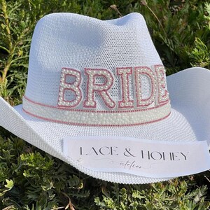 May include: White cowboy hat with a pink and white "BRIDE" embellishment with pearl accents. The hat has a pink ribbon and a white label that says "Lace & Honey atelier".