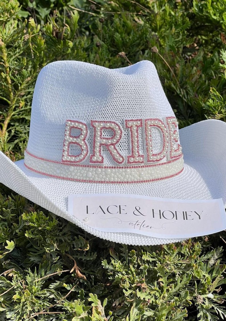 May include: White cowboy hat with a pink and white "BRIDE" embellishment with pearl accents. The hat has a pink ribbon and a white label that says "Lace & Honey atelier".