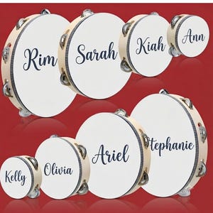 May include: A collection of seven round wooden tambourines of varying sizes, each featuring a white drumhead printed with a unique name in dark blue script. The names include Rim, Sarah, Kiah, Ann, Kelly, Olivia, Ariel, and Stephanie.