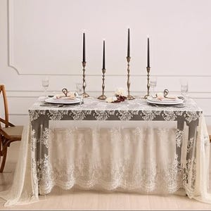 May include: A dining table set for a formal occasion. The table is covered with a gray tablecloth and a white lace overlay. Black candles in ornate holders, plates, and silverware are set for two. Wooden chairs flank the table.