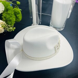 May include: A white cowboy hat with a pearl trim and a white satin ribbon. The hat is on a black surface.