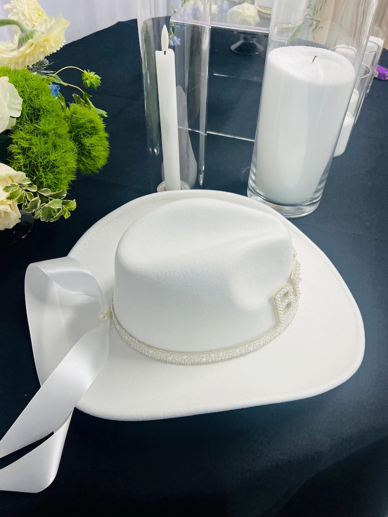 May include: A white cowboy hat with a pearl trim and a white satin ribbon. The hat is on a black surface.