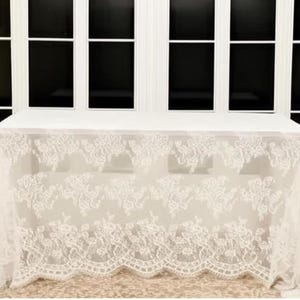 May include: A white rectangular table covered with a sheer, ivory lace tablecloth. The lace features a floral pattern along the edges, creating a delicate and elegant look. The table is set against a backdrop of large windows.