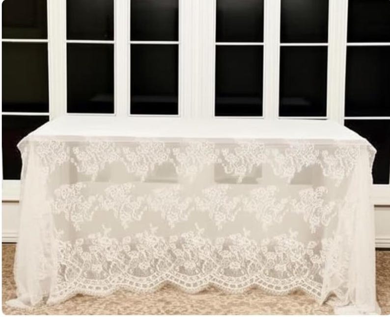 May include: A white rectangular table covered with a sheer, ivory lace tablecloth. The lace features a floral pattern along the edges, creating a delicate and elegant look. The table is set against a backdrop of large windows.