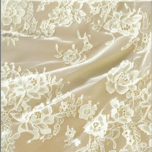 May include: Ivory lace fabric with a floral pattern. The delicate lace features detailed white flowers and leaves against a beige background. The fabric is draped, creating soft folds and shadows, highlighting the intricate design.