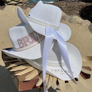May include: Two white cowboy hats with a white satin ribbon. The top hat has a white beaded trim and the bottom hat has a pink rhinestone "BRIDE" embellishment. The hats are sitting on a wooden surface with a brown straw-like material.