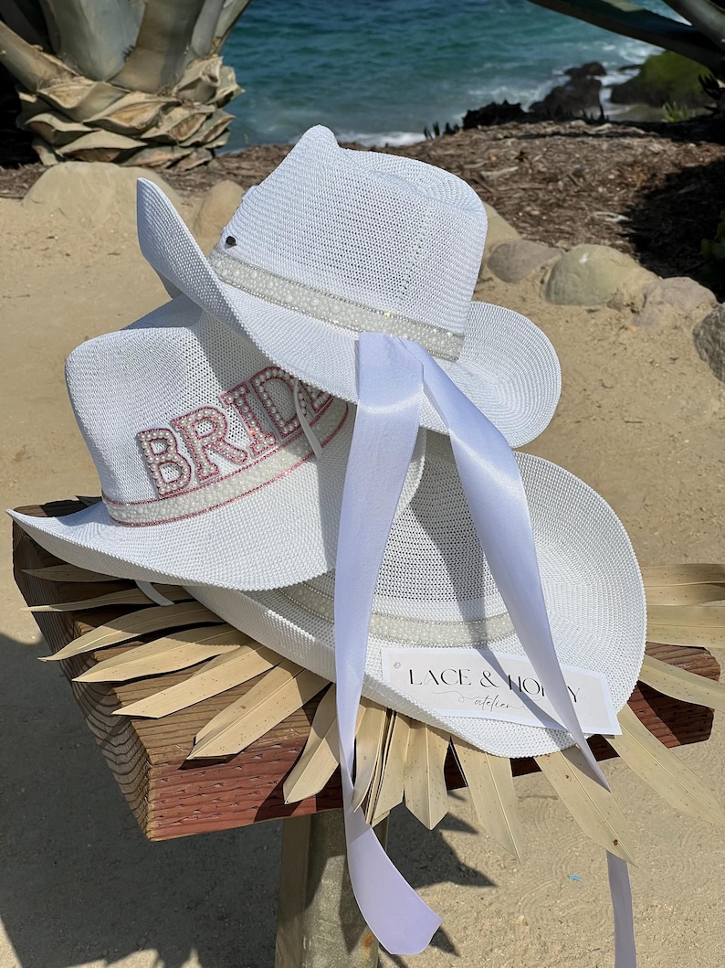 May include: Two white cowboy hats with a white satin ribbon. The top hat has a white beaded trim and the bottom hat has a pink rhinestone "BRIDE" embellishment. The hats are sitting on a wooden surface with a brown straw-like material.