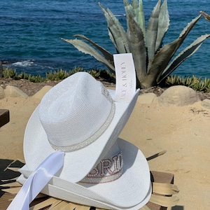 May include: White cowboy hat with a white satin ribbon and the word "Bride" in pink rhinestones. The hat is sitting on a wooden surface with a palm leaf.