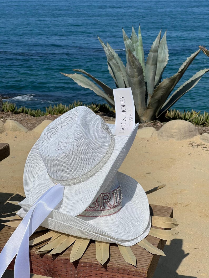 May include: White cowboy hat with a white satin ribbon and the word "Bride" in pink rhinestones. The hat is sitting on a wooden surface with a palm leaf.
