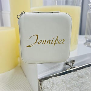 Pode incluir: Caixa quadrada branca com uma inscri&ccedil;&atilde;o dourada que diz "Jennifer". A caixa tem um fecho prateado e est&aacute; sobre uma superf&iacute;cie branca.