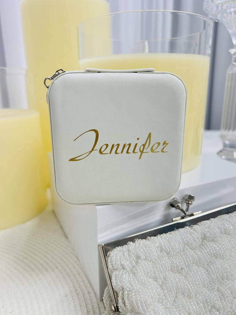 Pode incluir: Caixa quadrada branca com uma inscri&ccedil;&atilde;o dourada que diz "Jennifer". A caixa tem um fecho prateado e est&aacute; sobre uma superf&iacute;cie branca.